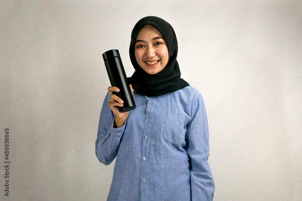 Beautiful young asian muslim woman smiling confidently, with hand holding black tumbler, hand holding black bottle, hand holding black glass, isolated on gray background, product advertising concept Beautiful young asian muslim woman smiling confidently, with hand holding black tumbler, hand holding black bottle, hand holding black glass, isolated on gray background, product advertising concept