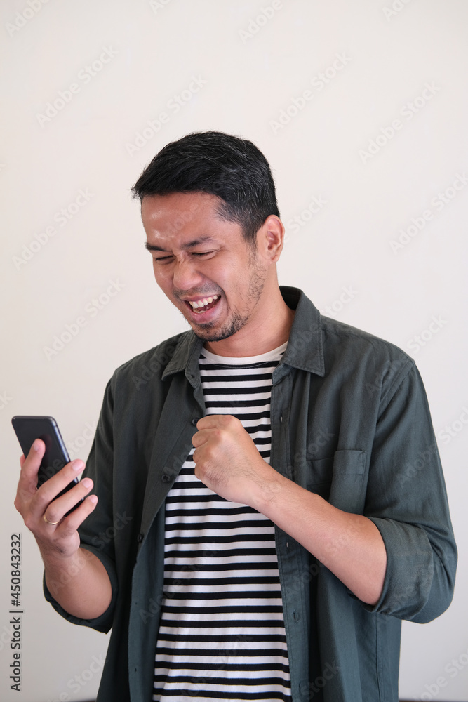 Adult Asian man showing excited gesture while looking to his mobile phone Adult Asian man showing excited gesture while looking to his mobile phone