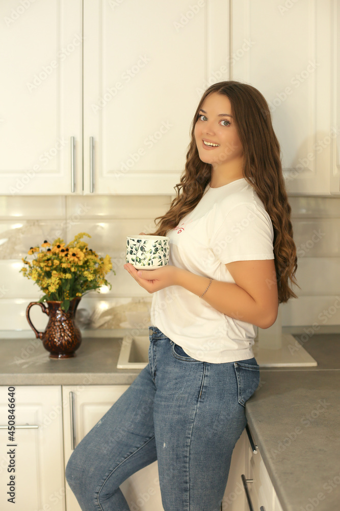 a young beautiful girl in a white T-shirt and jeans with dark long hair is standing in the kitchen holding a mug on the table there is a vase with wild flowers a young beautiful girl in a white T-shirt and jeans with dark long hair is standing in the kitchen holding a mug on the table there is a vase with wild flowers