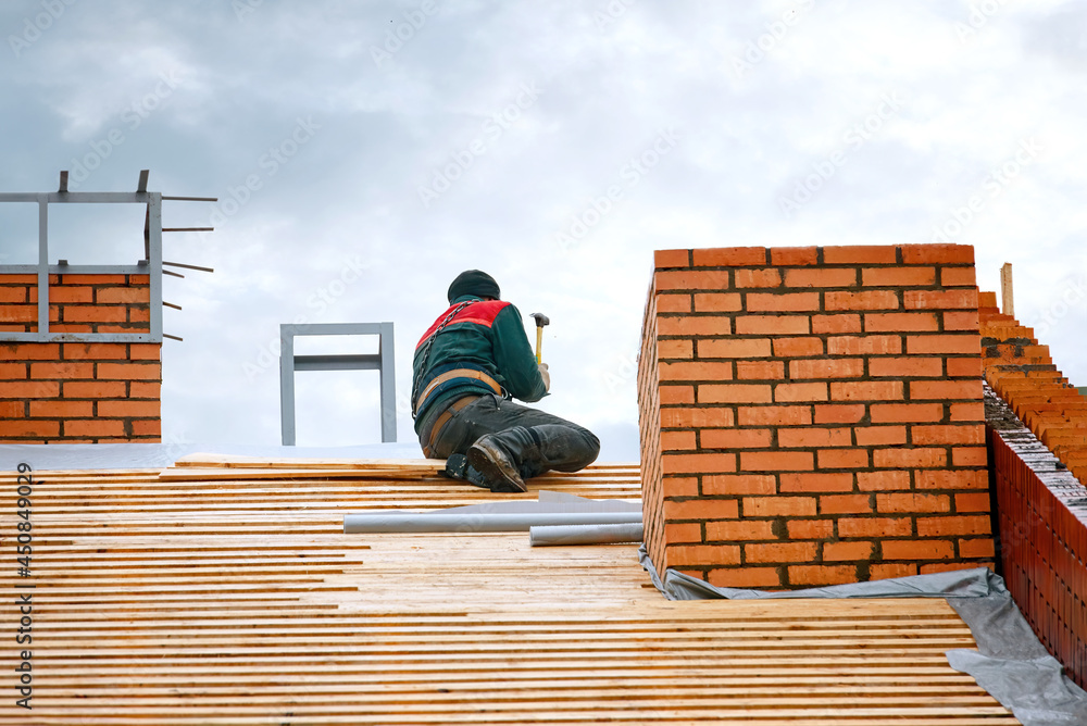 Worker hammers nails with hammer in wooden board on roof at height