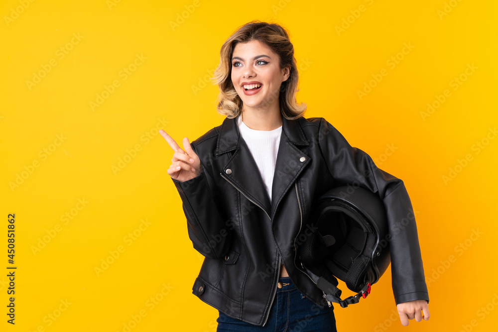 Teenager woman holding a motorcycle helmet isolated on yellow background surprised and pointing finger to the side Teenager woman holding a motorcycle helmet isolated on yellow background surprised and pointing finger to the side