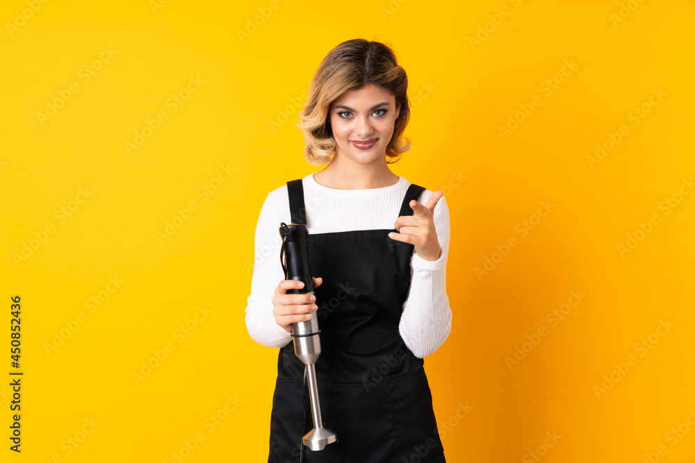 Girl using hand blender isolated on yellow background surprised and pointing front Girl using hand blender isolated on yellow background surprised and pointing front