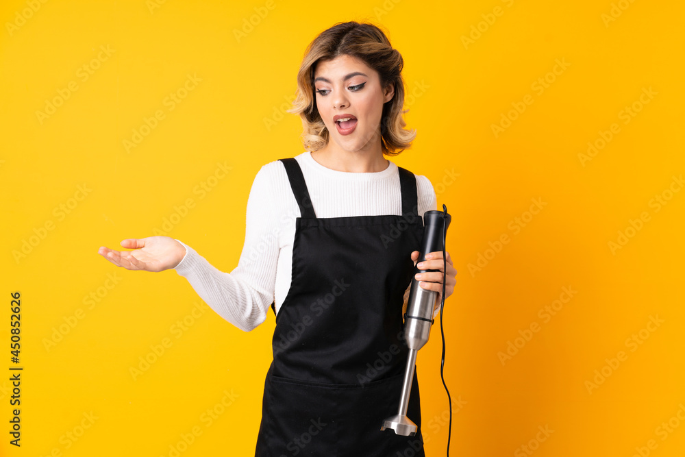 Girl using hand blender isolated on yellow background with surprise expression while looking side