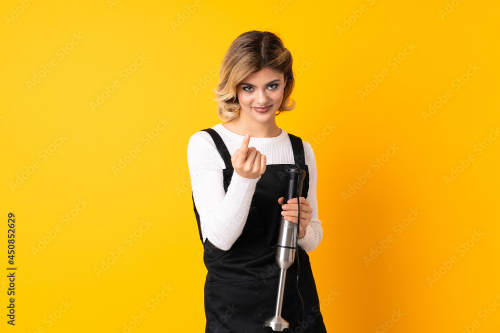 Girl using hand blender isolated on yellow background doing coming gesture