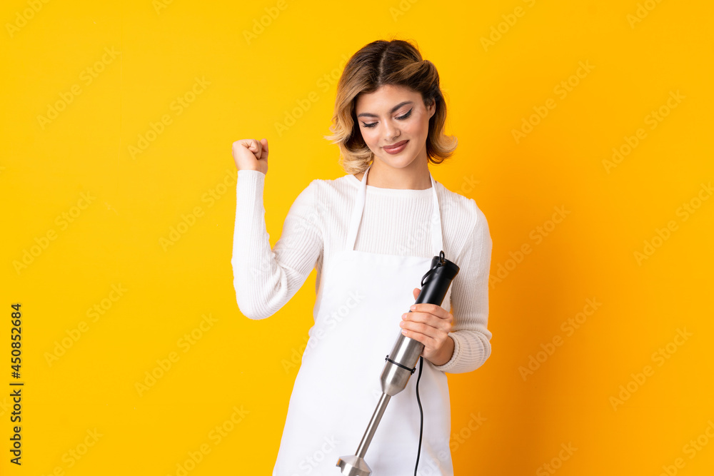 Girl using hand blender isolated on yellow background celebrating a victory