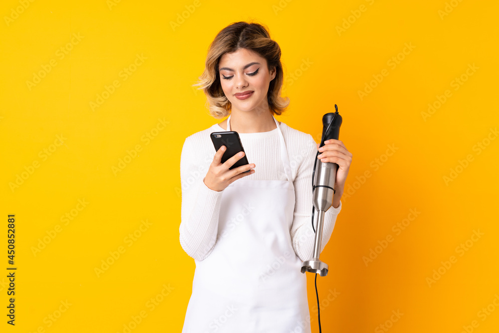 Girl using hand blender isolated on yellow background sending a message with the mobile Girl using hand blender isolated on yellow background sending a message with the mobile