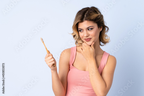 Teenager girl brushing her teeth isolated on blue background thinking an idea and looking side