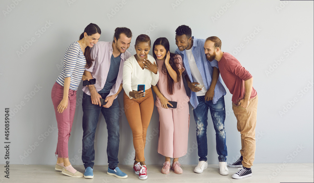 Fototapeta premium Modern technology. Young woman checks the camera on her new smartphone while taking a selfie with friends. Group of multiracial people with mobile phones in their hands standing against a light wall.