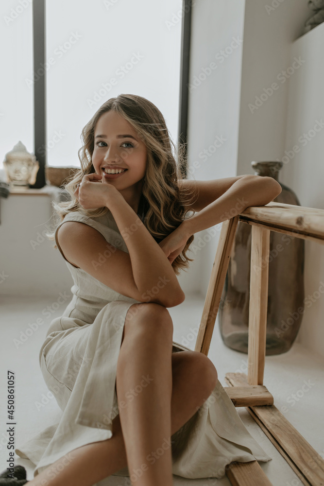 Happy cheerful woman in beige linen dress smiles gently, looks into camera, leans on wooden bench and sits on floor in white cozy bathroom. Happy cheerful woman in beige linen dress smiles gently, looks into camera, leans on wooden bench and sits on floor in white cozy bathroom.