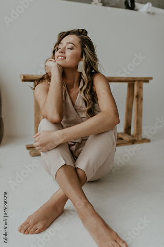 Calm blonde woman with wavy hair dressed in beige stylish jumpsuit smiles gently and sits on floor in white room.
