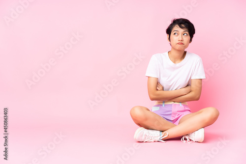 Young Vietnamese woman with short hair sitting on the floor over isolated pink background making doubts gesture while lifting the shoulders