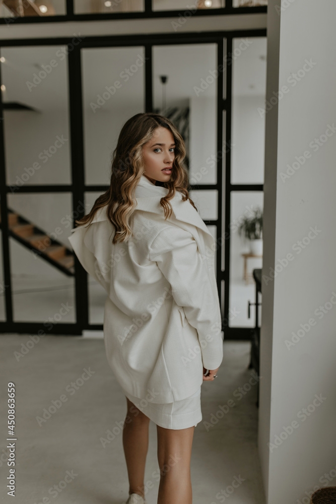 Pretty woman in white skirt, oversized stylish jacket looks back and poses in beautiful cozy light living room. Pretty woman in white skirt, oversized stylish jacket looks back and poses in beautiful cozy light living room.