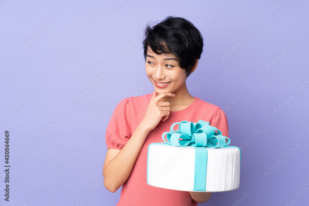 Young Vietnamese woman with short hair holding a big cake over isolated purple background thinking an idea and looking side Young Vietnamese woman with short hair holding a big cake over isolated purple background thinking an idea and looking side