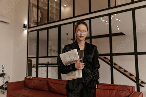 Calm serious woman in black silk home suit looks into camera and poses in cozy living room. Charming lady in eyeglasses holds newspaper and laptop.