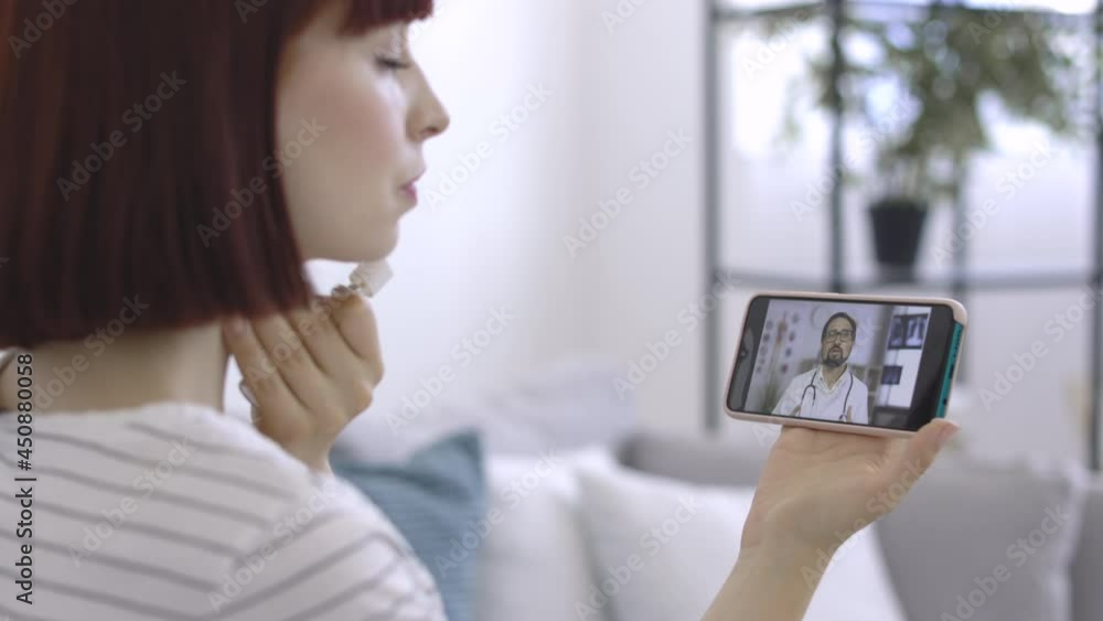 Online drug prescribing. Pretty suffering sick woman with a sore throat, using oral spray for treatment, having a video call from home, listening to the male doctor, giving recommendations Online drug prescribing. Pretty suffering sick woman with a sore throat, using oral spray for treatment, having a video call from home, listening to the male doctor, giving recommendations