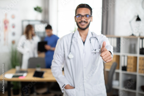 Medical education and healthcare. Blur view with copy space of young smiling male medical student showing thumb up to camera while standing in modern study room with groupmates on background