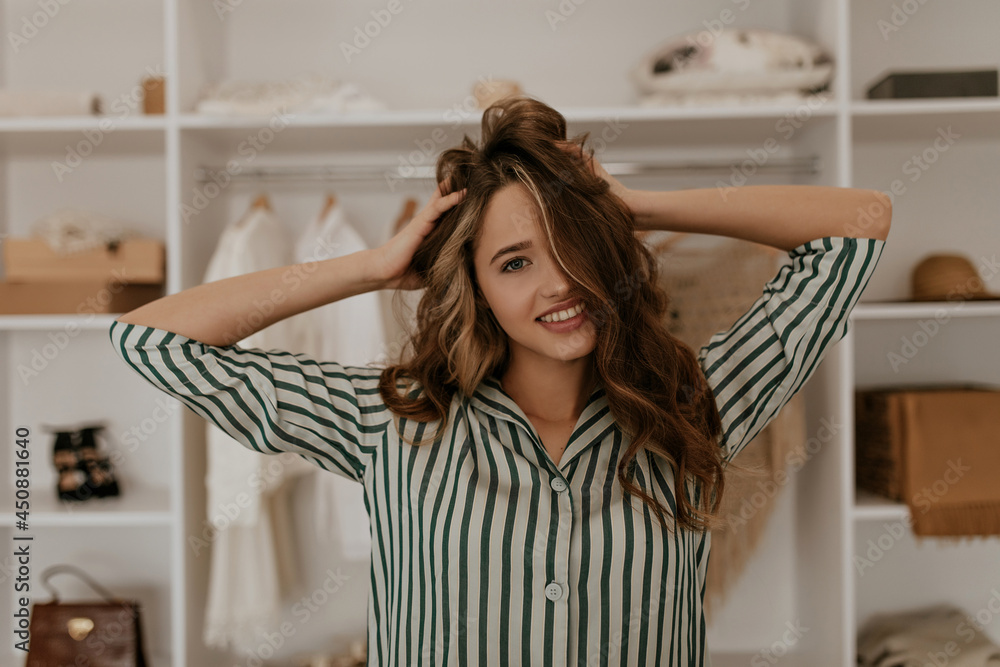 Portrait of curly charming girl ruffles hair in dressing room. Cool young woman in silk striped shirt looks into camera and smiles. Portrait of curly charming girl ruffles hair in dressing room. Cool young woman in silk striped shirt looks into camera and smiles.