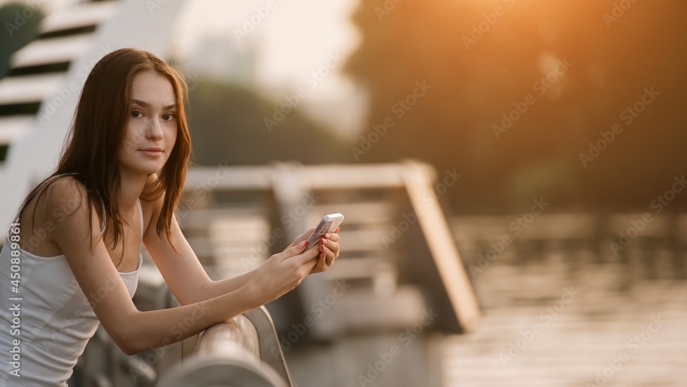 Beautiful young girl walking on a park and chatting on a mobile phone during sunset         