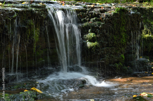Fototapeta Naklejka Na Ścianę i Meble -  A small waterfall formed by a small forest river, inside a shaded ravine, on a sunny day.