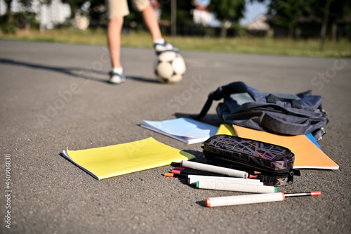 Scattered school supplies and workbooks falling out of an open backpack, lie on the asphalt of the schoolyard against the background of a boy's feet on a soccer ball.