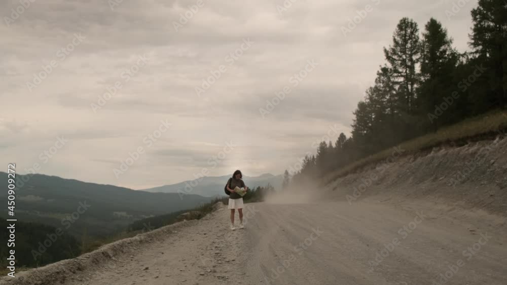 The girl is hitchhiking alone. He walks along the road with a paper map and tries to stop passing cars. Beautiful landscape: dusty road, forest, mountains. The girl is hitchhiking alone. He walks along the road with a paper map and tries to stop passing cars. Beautiful landscape: dusty road, forest, mountains.