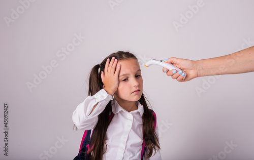 a sick schoolgirl in uniform and with a backpack measures the temperature with an infrared thermometer on a white background with a place for text