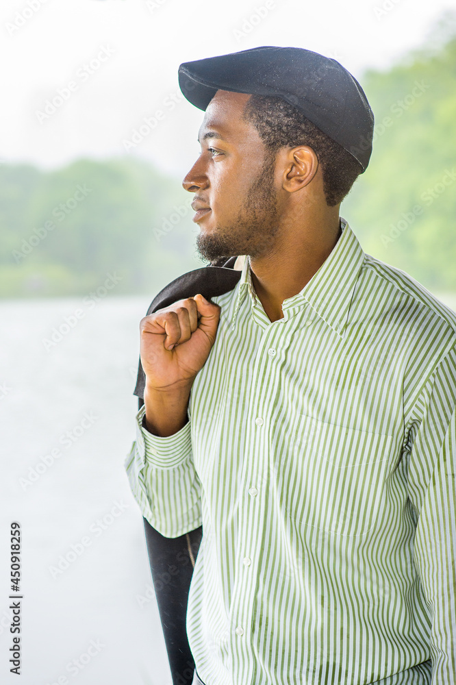 Fototapeta premium Dressing in a long sleeves shirt, a ivy cap, taking off his jacket, a young handsome black guy is standing by a lake in a raining, foggy day, smilingly looking at you..