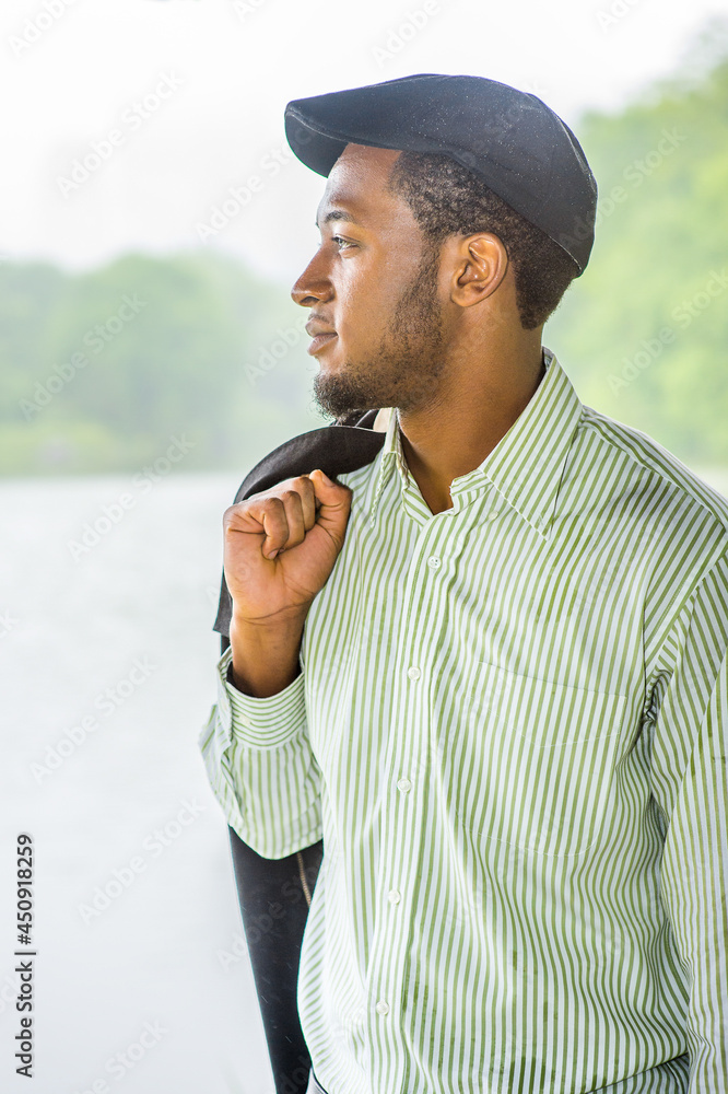Dressing in a long sleeves shirt, a ivy cap, taking off his jacket,  a young handsome black guy is standing by a lake in a raining, foggy day, smilingly looking at you..