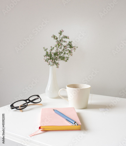 Close up of pink disoma flowers in small vase, glasses and tea cup on white table with pink journal and pen in foreground (selective focus)