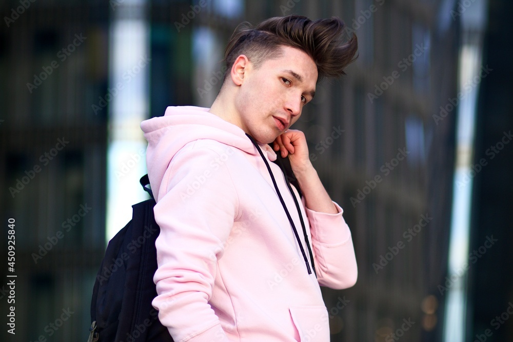 Portrait of young man handsome pretty stylish guy with long hair university or college student in pink hoodie and backpack looking at camera outdoors. Stock Adobe Stock
