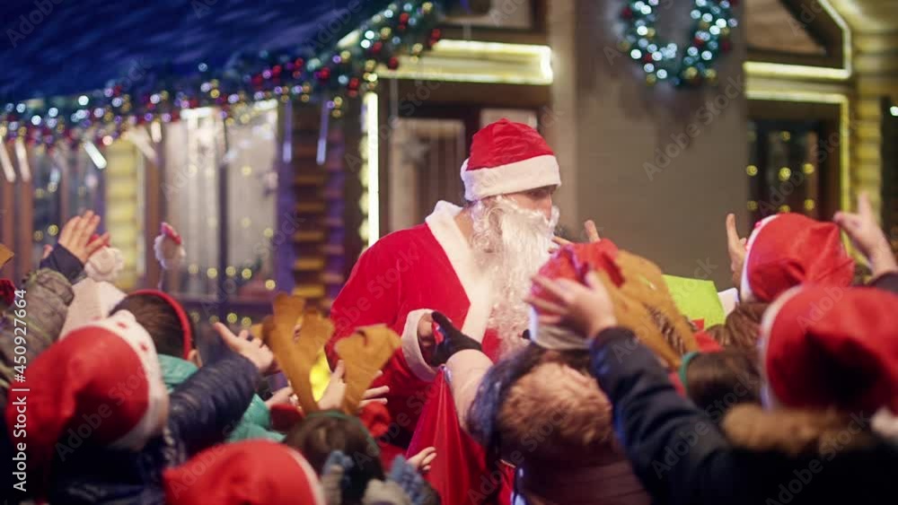 Santa Claus gives wrapped gift boxes to cute little children . Two Santa clauses helping each other to give gifts to queue of children . Many kids standing and waiting in line queue for a gift . Santa Claus gives wrapped gift boxes to cute little children . Two Santa clauses helping each other to give gifts to queue of children . Many kids standing and waiting in line queue for a gift .