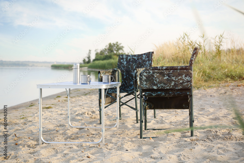 Camouflage fishing chairs and table with metal cups on sandy beach near river
