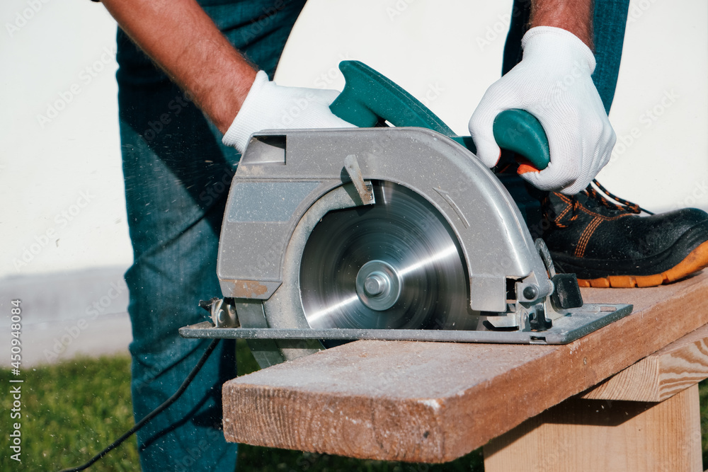 A male joiner works with a hand-held circular saw. A builder is sawing a board at the construction site of a new house.