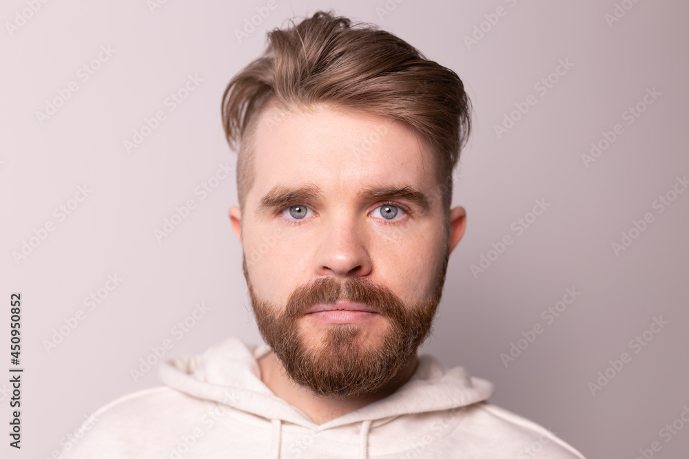 Obraz premium Portrait of young male with beard and mustache and trendy hairdo. Wears casual hoodie, has serious expression in studio against white background