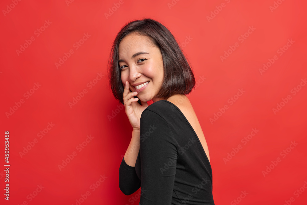 Sideways shot of pretty Asian woman smiles gently has shy happy expression wears black dress expresses positive authentic emotions isolated over vivid red background. Lovely brunette ethnic female Sideways shot of pretty Asian woman smiles gently has shy happy expression wears black dress expresses positive authentic emotions isolated over vivid red background. Lovely brunette ethnic female
