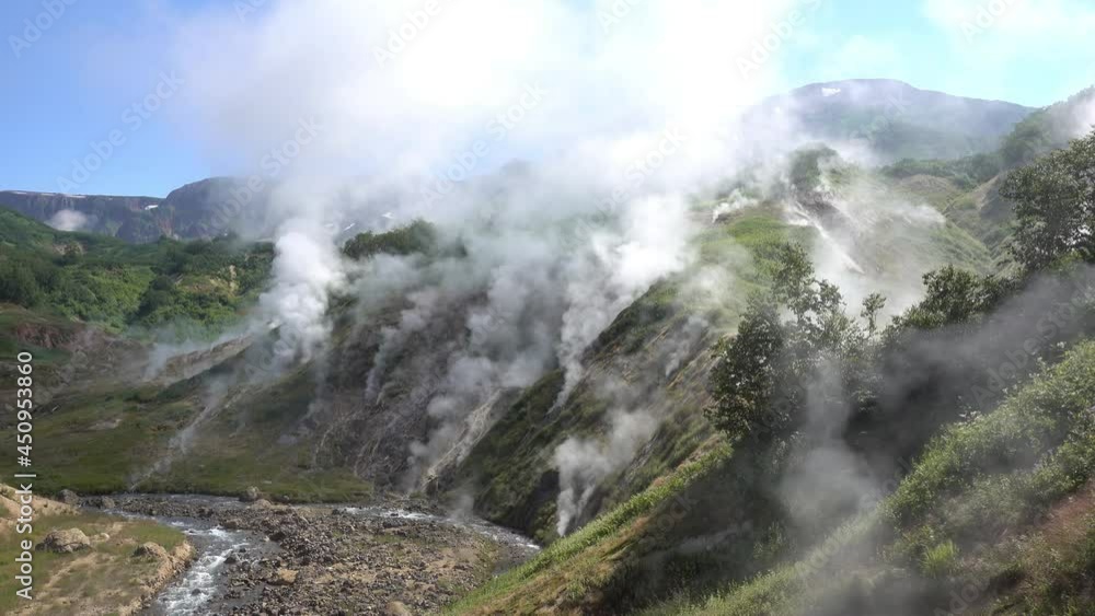 A Unique Valley Of Geysers. Kamchatka. Steam from hot springs rises into the blue sky above the mountain slopes. The river flows along a rocky bed. Green vegetation around A Unique Valley Of Geysers. Kamchatka. Steam from hot springs rises into the blue sky above the mountain slopes. The river flows along a rocky bed. Green vegetation around