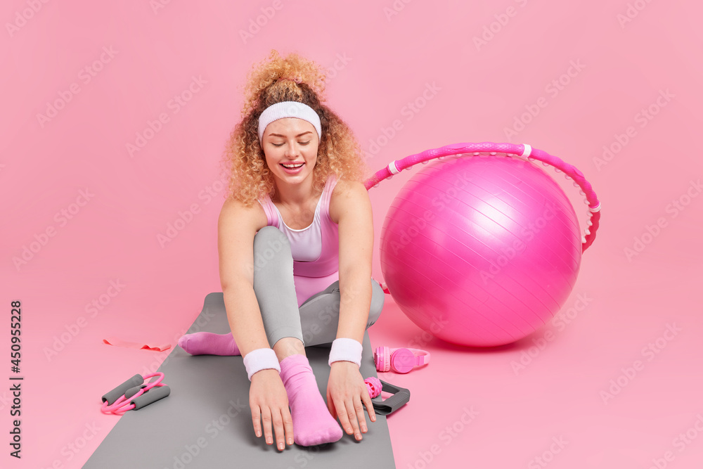 Curly haired young active woman leads sporty lifestyle stretches on fitness mat leans to legs wears headband sportswear uses fitness ball hula hoop elastic band for workout has training at home Curly haired young active woman leads sporty lifestyle stretches on fitness mat leans to legs wears headband sportswear uses fitness ball hula hoop elastic band for workout has training at home