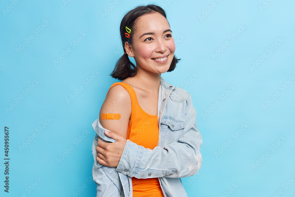 Pleased teenage girl with eastern appearance has two pony tails smiles gently happy to get vaccine against coronavirus dressed in orange dress and denim jacket isolated over blue background. Pleased teenage girl with eastern appearance has two pony tails smiles gently happy to get vaccine against coronavirus dressed in orange dress and denim jacket isolated over blue background.