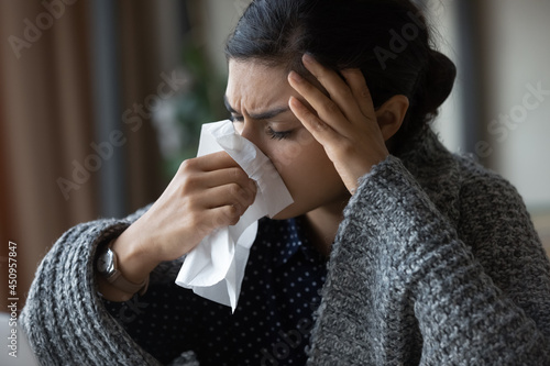 Unhealthy young Indian woman covered in blanket blow nose suffer from flu or cold. Unwell millennial biracial female feel sick ill at work, struggle with corona virus covid-19. Healthcare concept.