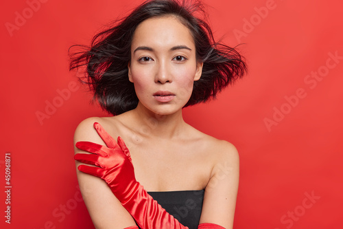 Gorgeous young Asian woman with dark hair floating in air looks seriously at camera wears elegnat black dress long gloves stands bare shoulders has healthy skin isolated over red background. photo