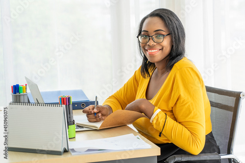 South American African woman wearing a yellow shirt works as a secretary writing planners, diaries and appointments at her desk.  