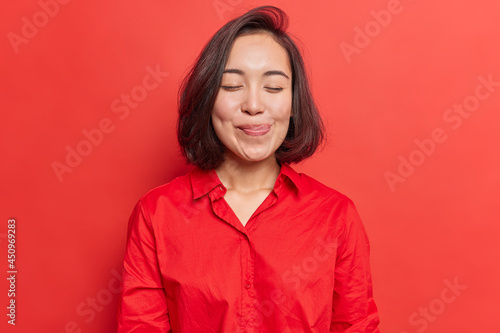 Pretty brunette young Asian woman closes eyes licks lipsfrom temptation to taste something delicious shows tongue imagines eating delicious food wears shirt isolated over vivid red background photo