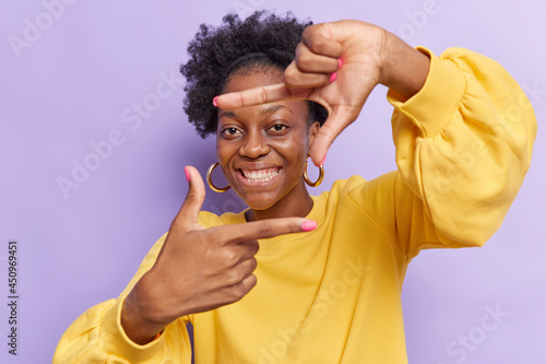 Happy ambitious dark skinned woman aims only success makes hand frames searches perfect angle smiles broadly wears yellow sweater isolated over purple background gazes at camera through hands