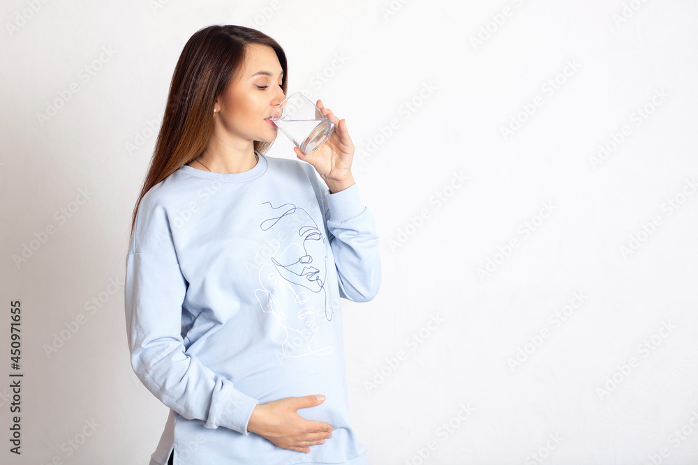 A beautiful happy pregnant woman drinks water from a glass glass. White background, space for text. Health care. Proper nutrition. A beautiful happy pregnant woman drinks water from a glass glass. White background, space for text. Health care. Proper nutrition.