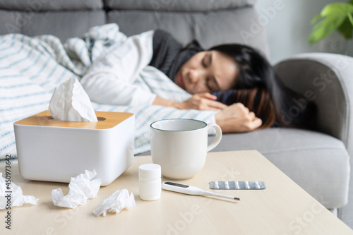 Asian woman suffering from covid-19 symptoms have to be on self-isolation at home. Girl has a fever lying on couch with tissue, pill and a glass of water on desk. Cold, flu and health problems concept