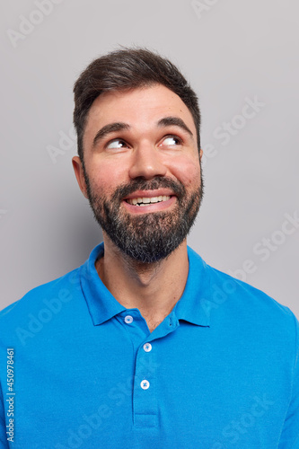 Vertical shot of dreamy satisfied bearded adult man look up considers something recalls pleasant moment in life smiles and shows teeth wears casual blue t shirt isolated over grey background.