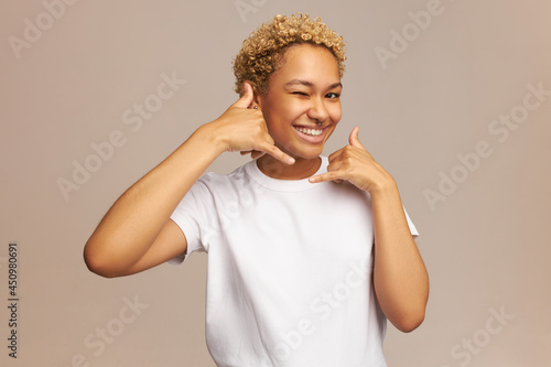 Studio shot of good-looking winking young joyful African girl with blonde curly haircut and nose piercing, smiling showing healthy white teeth, making call me later gesture with phone shaped hand