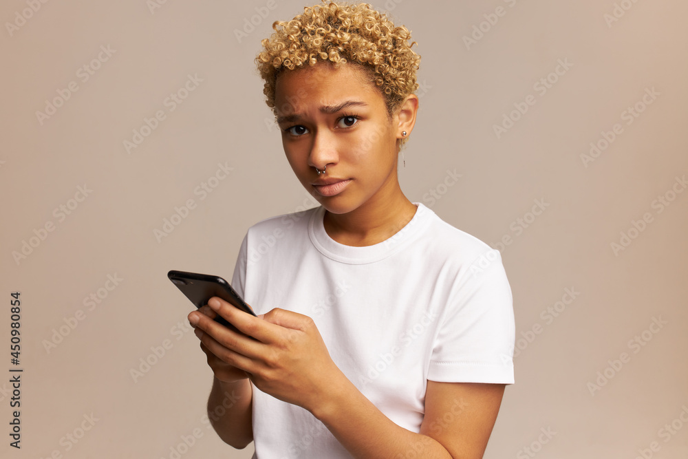 Upset African woman wearing white T-shirt and blonde short hair, isolated against pink background, looking at camera with sad face expression, feeling unhappy, displeased, holding mobile phone Upset African woman wearing white T-shirt and blonde short hair, isolated against pink background, looking at camera with sad face expression, feeling unhappy, displeased, holding mobile phone