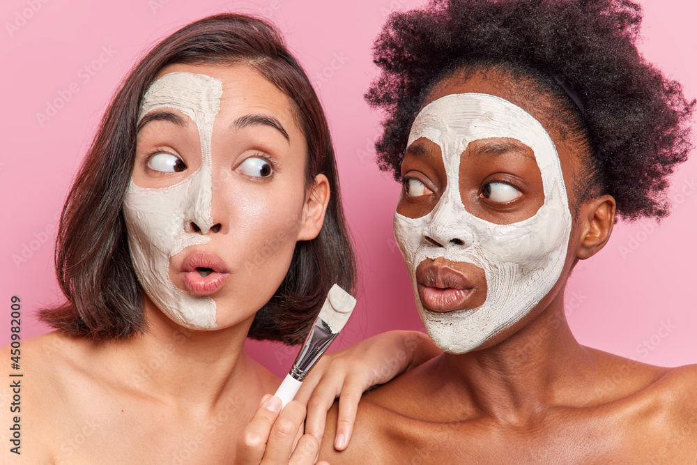 Surprised mixed race friendly women apply nourishing face masks with cosmetic brush look shocked at each other stand shirtless against pink background. Skin care beauty and wellness concept. Surprised mixed race friendly women apply nourishing face masks with cosmetic brush look shocked at each other stand shirtless against pink background. Skin care beauty and wellness concept.