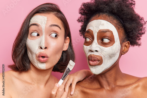 Surprised mixed race friendly women apply nourishing face masks with cosmetic brush look shocked at each other stand shirtless against pink background. Skin care beauty and wellness concept.